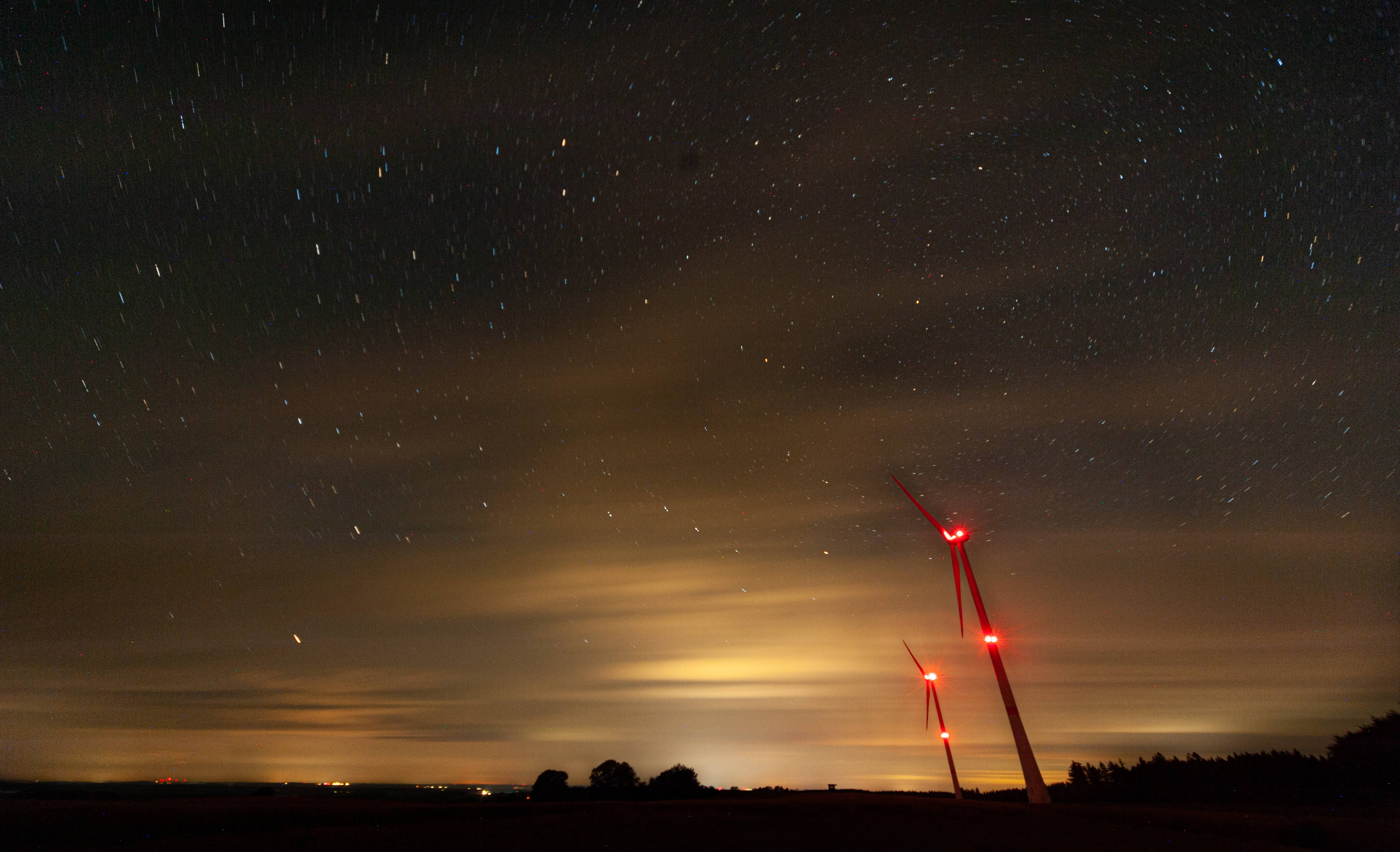 Two motionless wind turbines in a cloudy night. They are reddened by their anti-collision lights in the middle and on the to p. In the background, there are clouds lit up in yellow-orangy colors by the village below. Above the clouds, a sky full of stars is recognizable.
