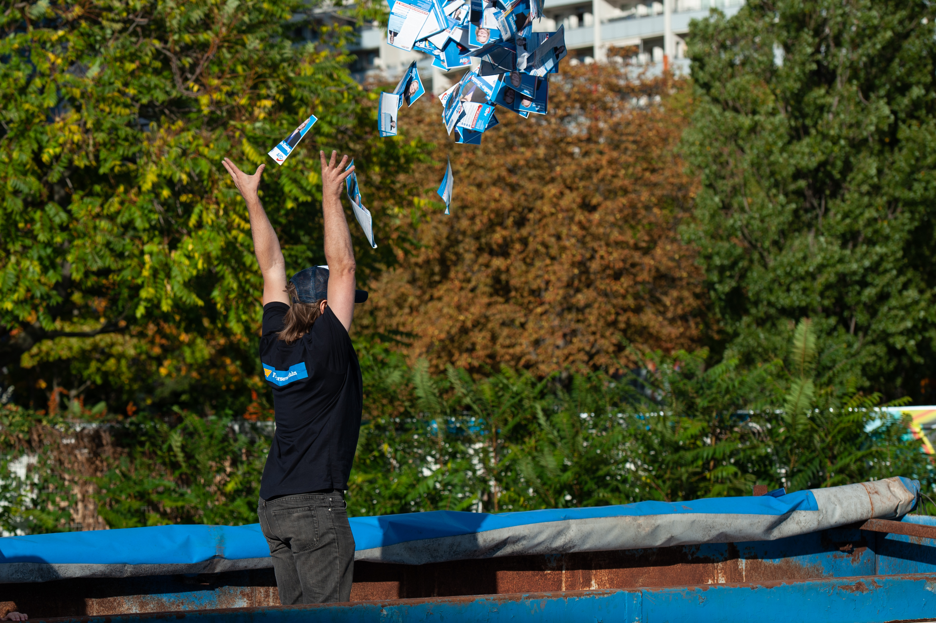 The person is now standing inside a larger waste container filled up high with flyers. They are throwing a bunch of them into the air. The photo is taken from behind their right side in a wide perspective, they are visible from the knees up to the head. I the background, there are some trees and a multi-story apartment building.